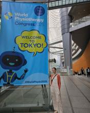Cara Whalen Smith stands smiling in the Tokyo International Forum besides a large banner that reads 'World Physiotherapy Congress 2025: Welcome to Tokyo!'